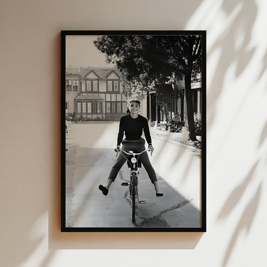 A black and white photograph of a person riding a bicycle on a street, with a building and trees in the background.