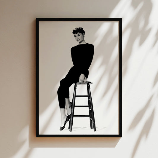 A black and white photograph of a woman sitting on a stool, wearing a long-sleeved top and pants.