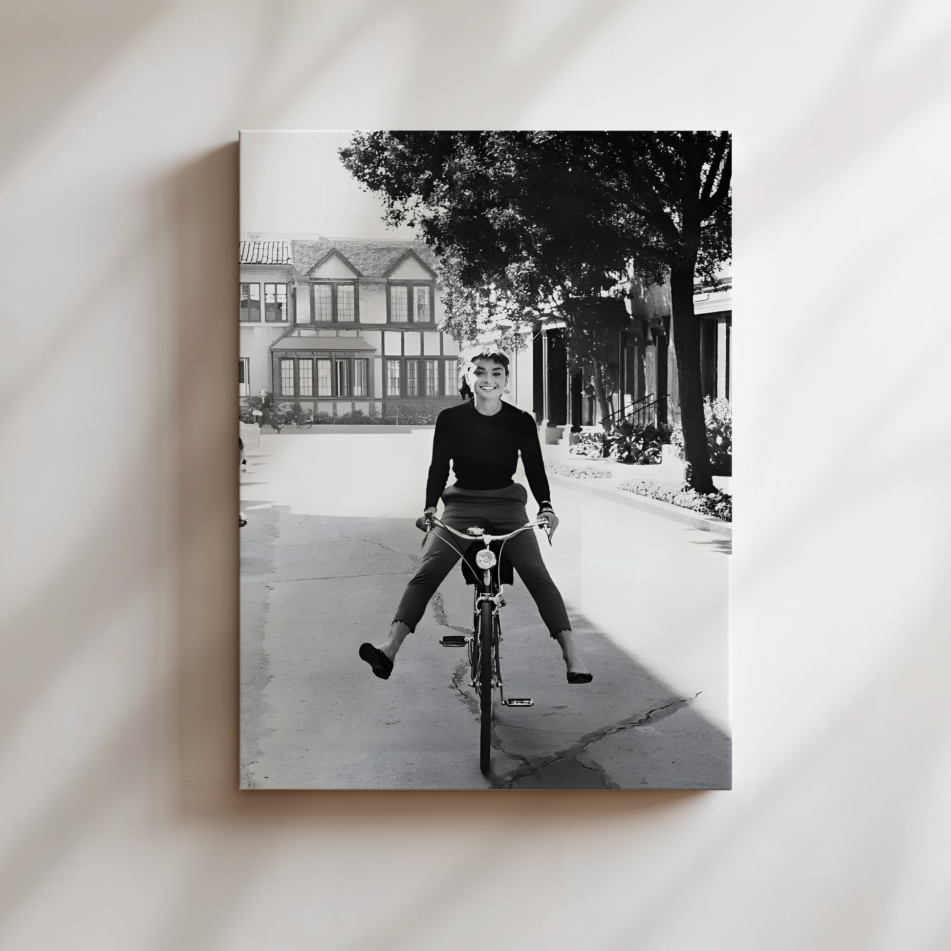 A black and white photograph of a person riding a bicycle on a street, with a house and trees in the background.
