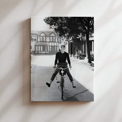 A black and white photograph of a person riding a bicycle on a street, with a house and trees in the background.