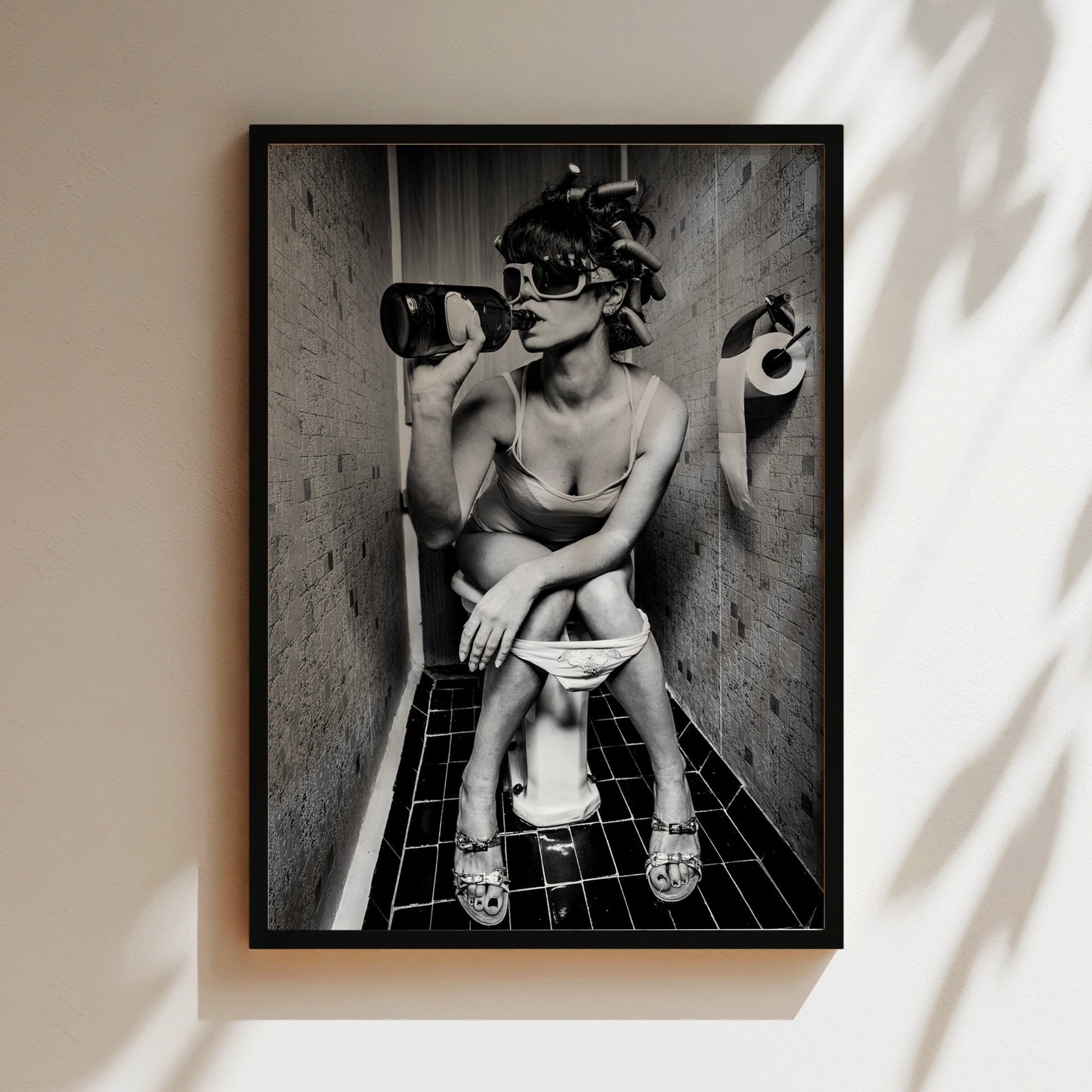 A black and white image of a woman sitting on a toilet in a bathroom, wearing sunglasses and holding a bottle.