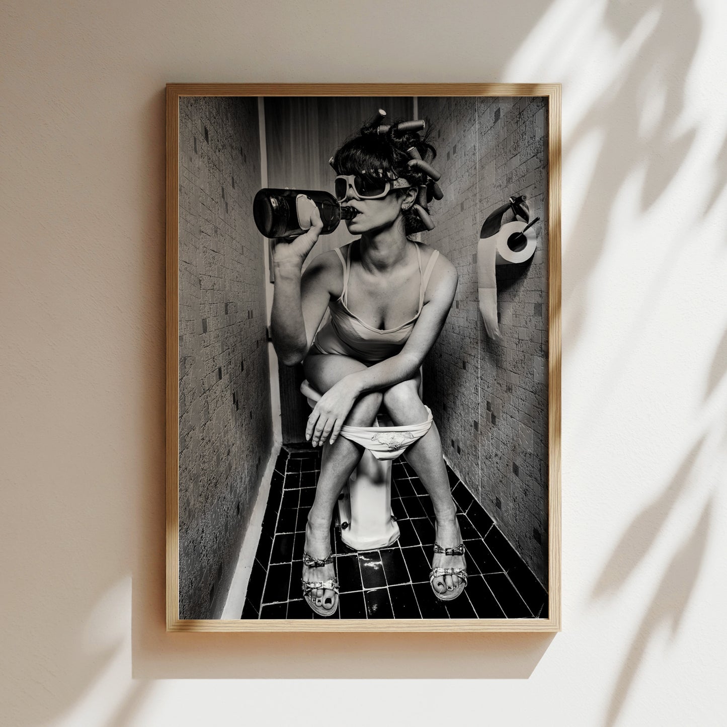 A black and white photograph of a woman sitting on a toilet in a bathroom, wearing sunglasses and holding a bottle.