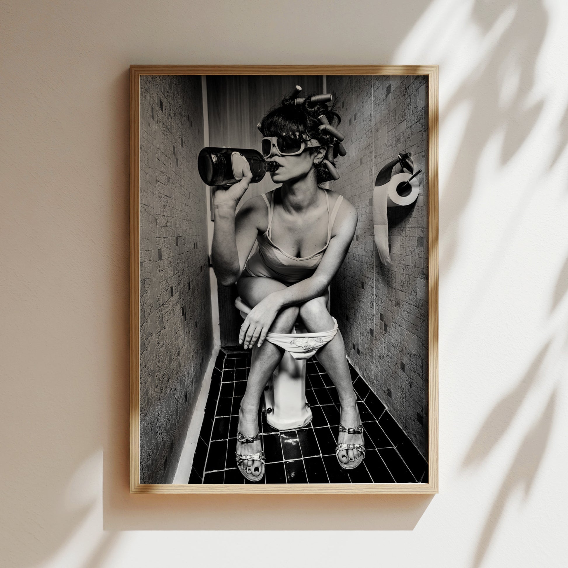 A black and white photograph of a woman sitting on a toilet in a bathroom, wearing sunglasses and holding a bottle.