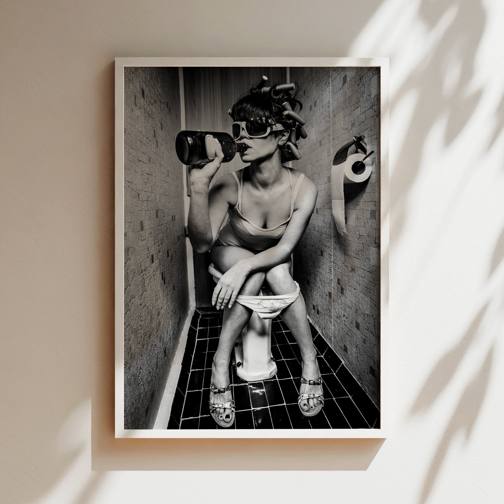 A black and white photograph of a woman sitting on a toilet in a bathroom, wearing a tank top and sunglasses.