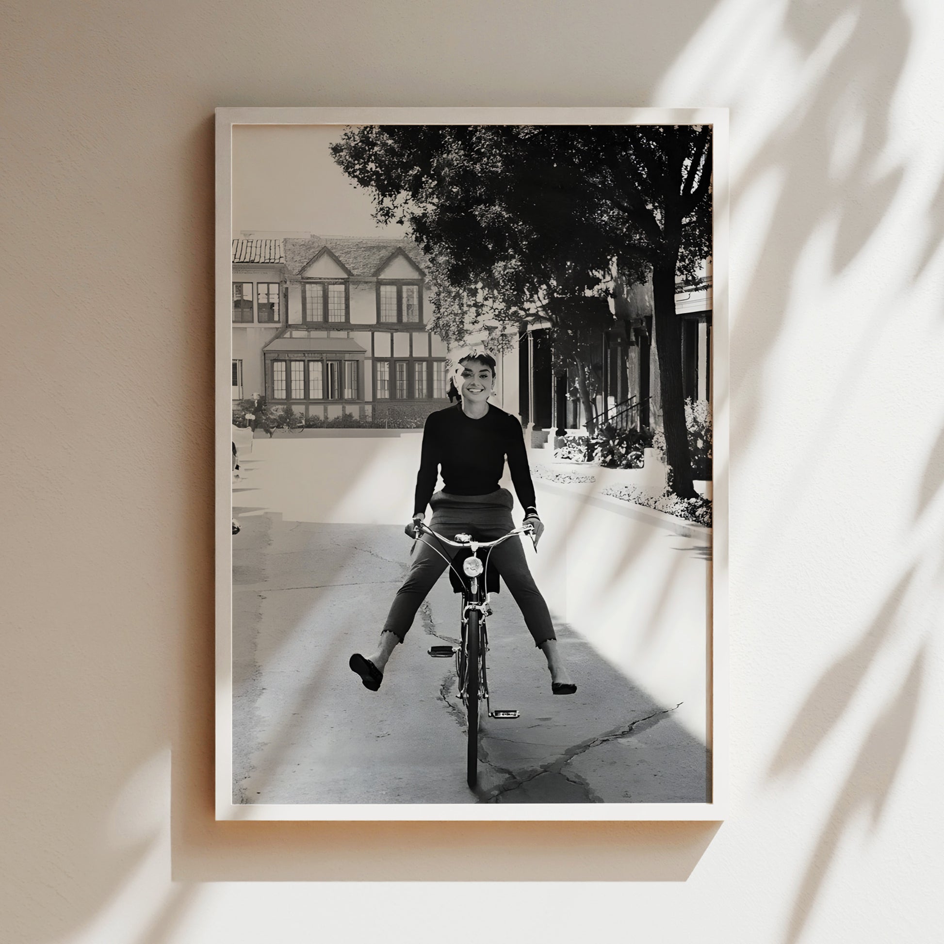A black and white photograph of a person riding a bicycle on a street, with a house and trees in the background.